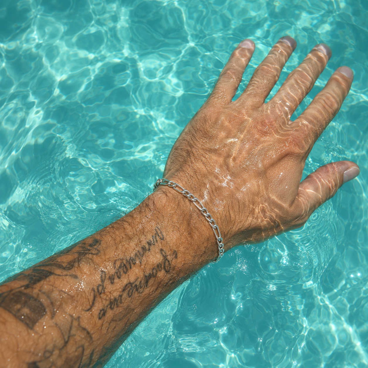 A close-up of a tattooed hand resting in clear turquoise pool water, wearing a silver chain bracelet.