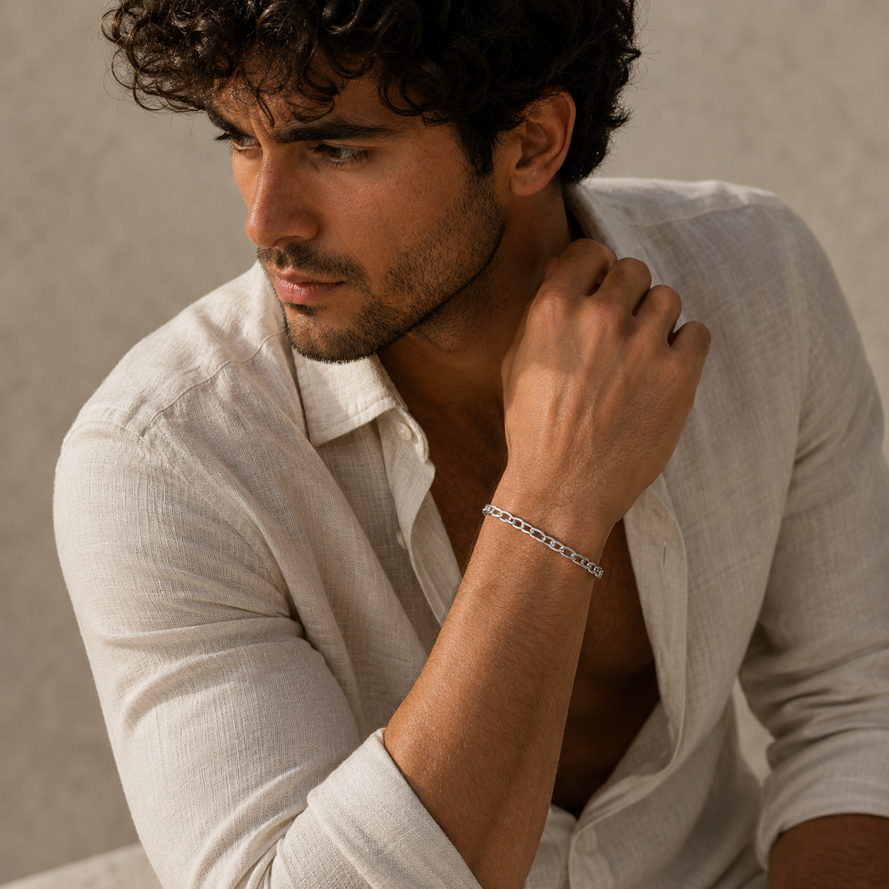 A close-up lifestyle shot of a man wearing a silver chain link bracelet on his wrist, dressed in a light beige linen shirt against a neutral textured background.