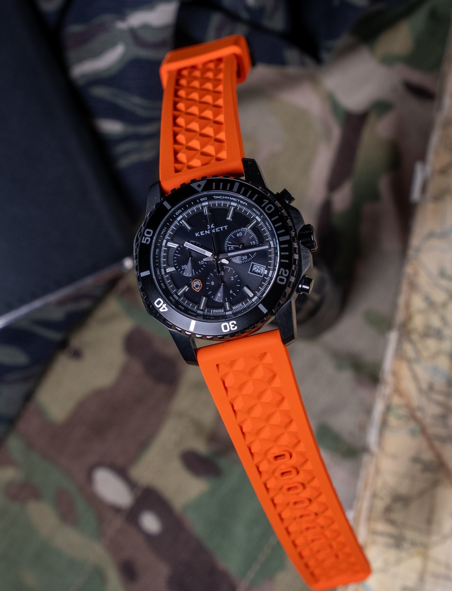 Kennett x Ollie Ollerton Solaforce watch with orange rubber strap laid flat on camouflage military fabric background